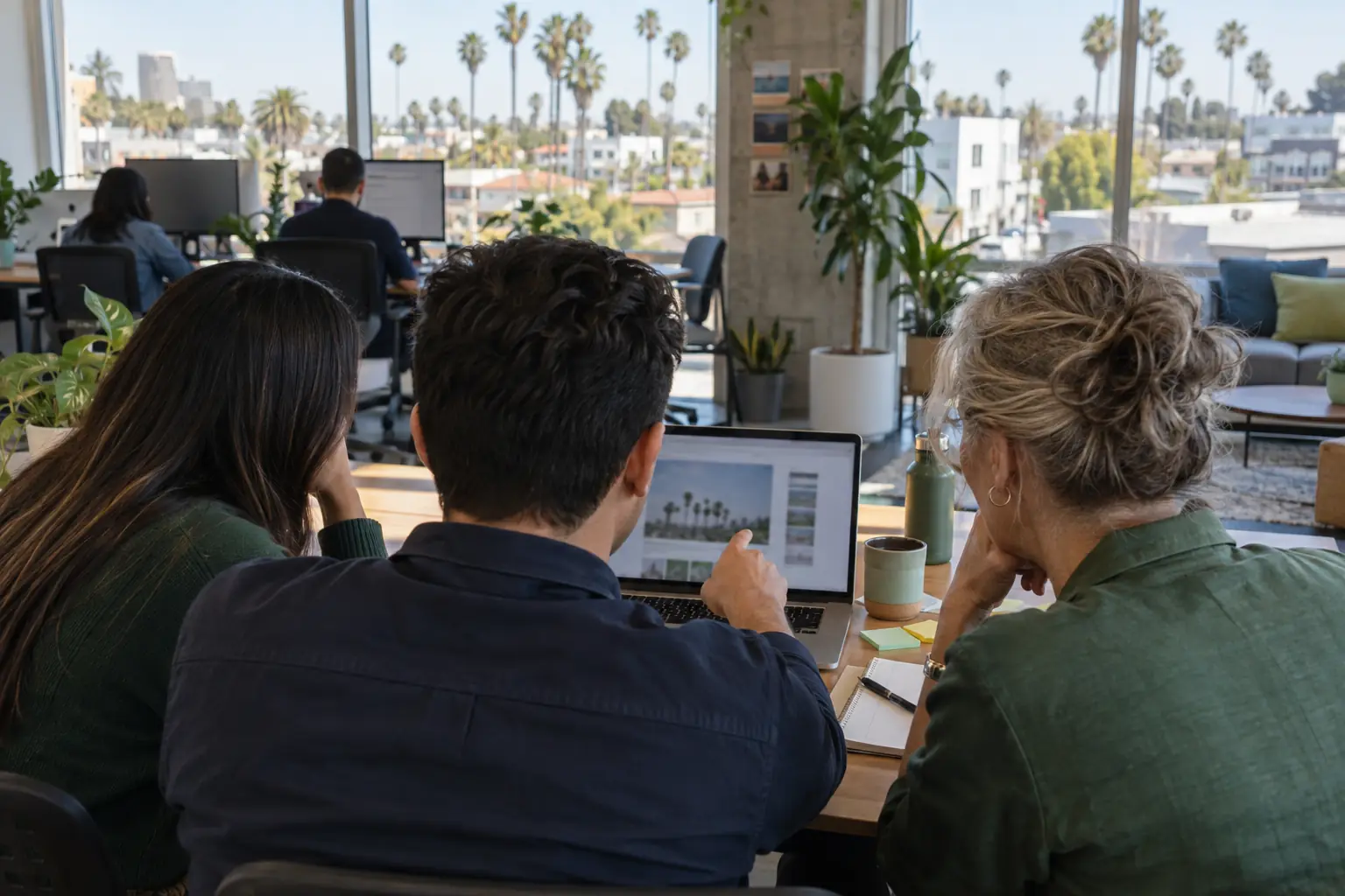Three coworkers gathered around one laptop at a sunny shared table, trying out a new tool together