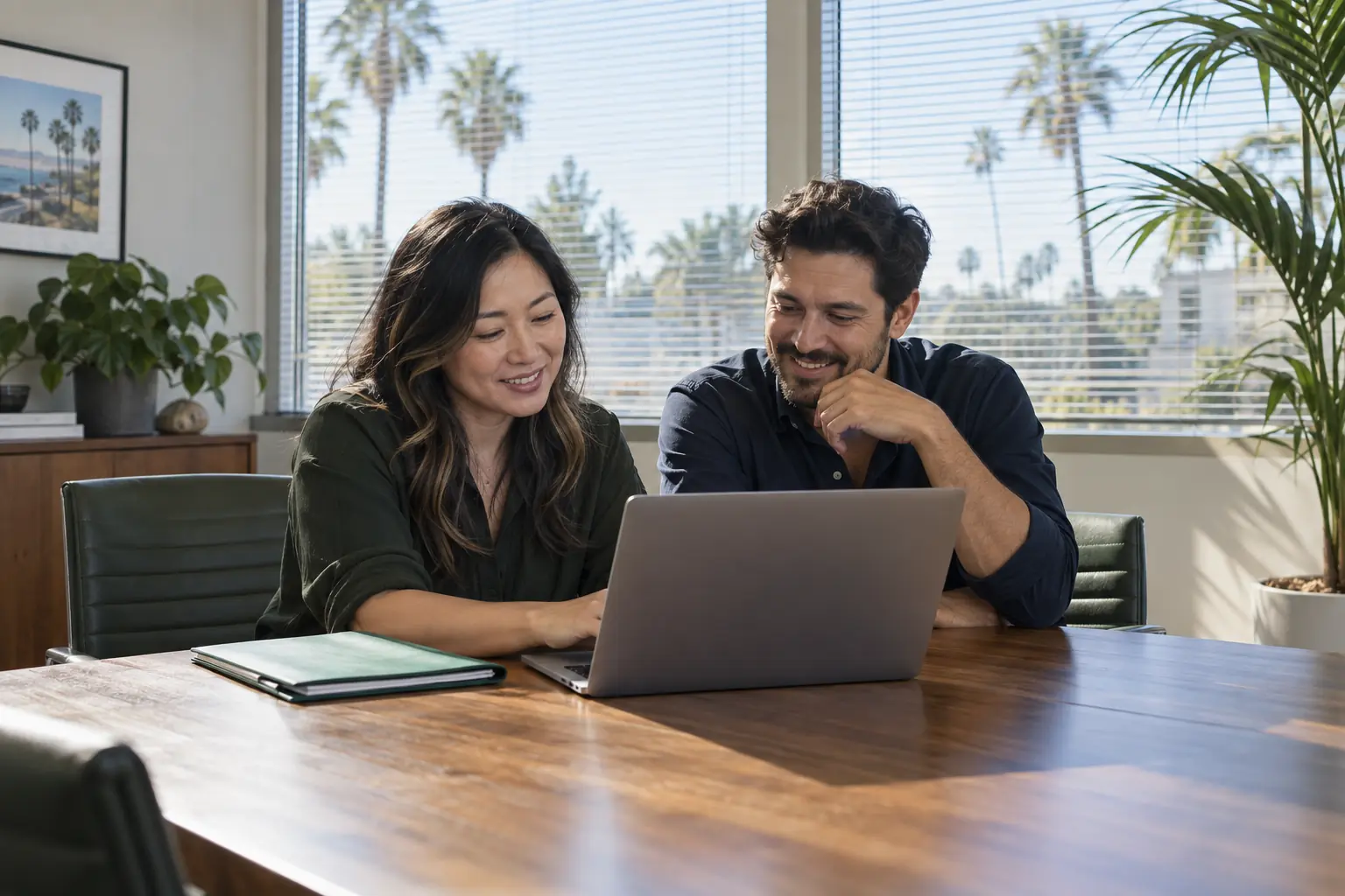 Two colleagues smiling as they review a laptop together at a conference table in a sunny office